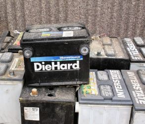 Stacked used car batteries, including a black DieHard battery, in a cluttered outdoor area