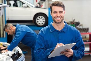 Mechanic holding a clipboard in an auto repair shop, with a worker fixing a car in the background