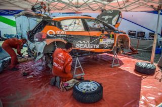 Rally car in a pit tent with mechanics repairing it, orange and black livery, wheels removed on red floor.