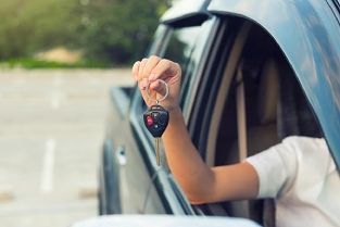 Car window view of a hand holding up car keys beside a blue vehicle