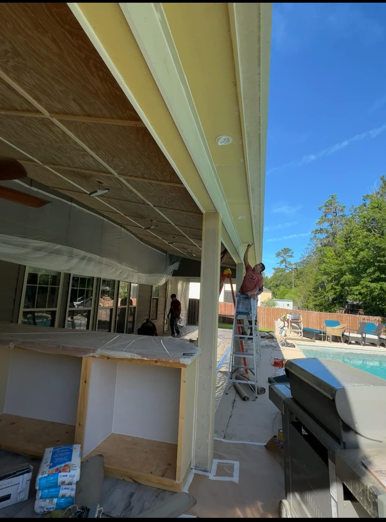Person on ladder painting exterior of a building beside a pool under a blue sky