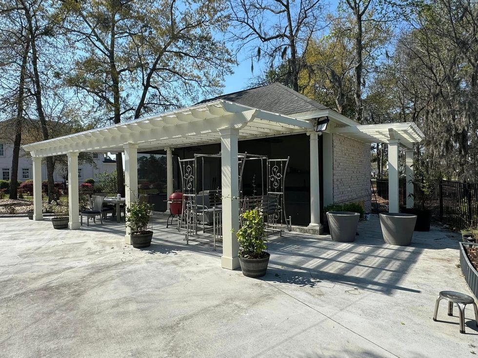 White patio with pergola, potted plants, and outdoor seating beside a dark building in a wooded area