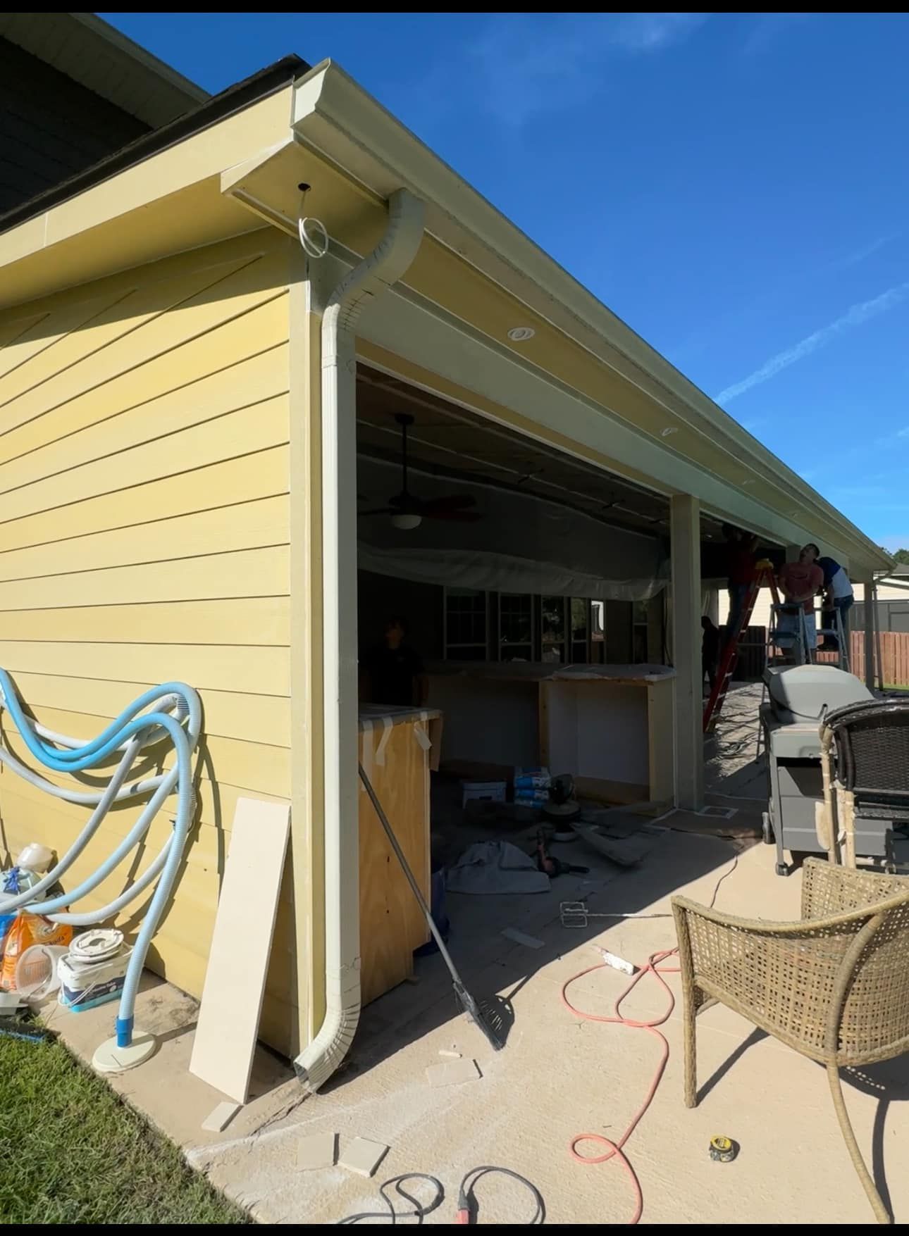 Yellow house exterior with open garage, hoses, and patio furniture in a sunny yard