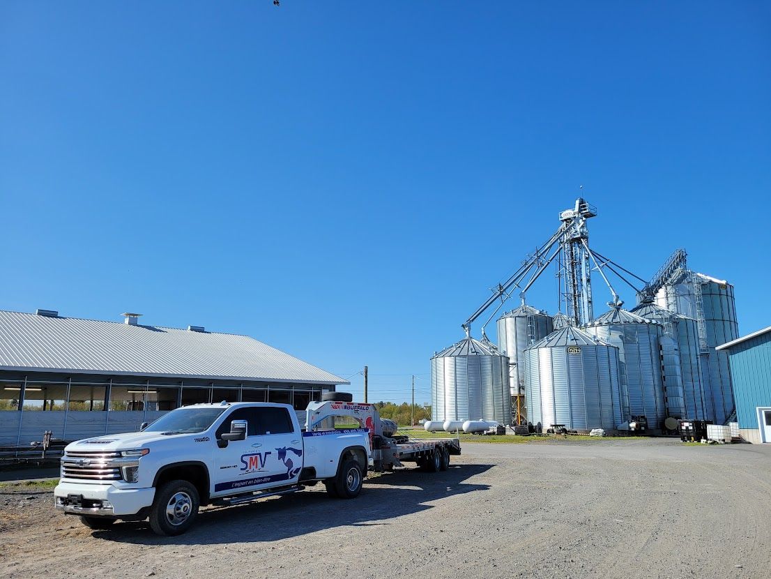 White pickup truck with a trailer parked in front of grain silos on a sunny day. Blue sky, gravel ground, and a light-colored building.