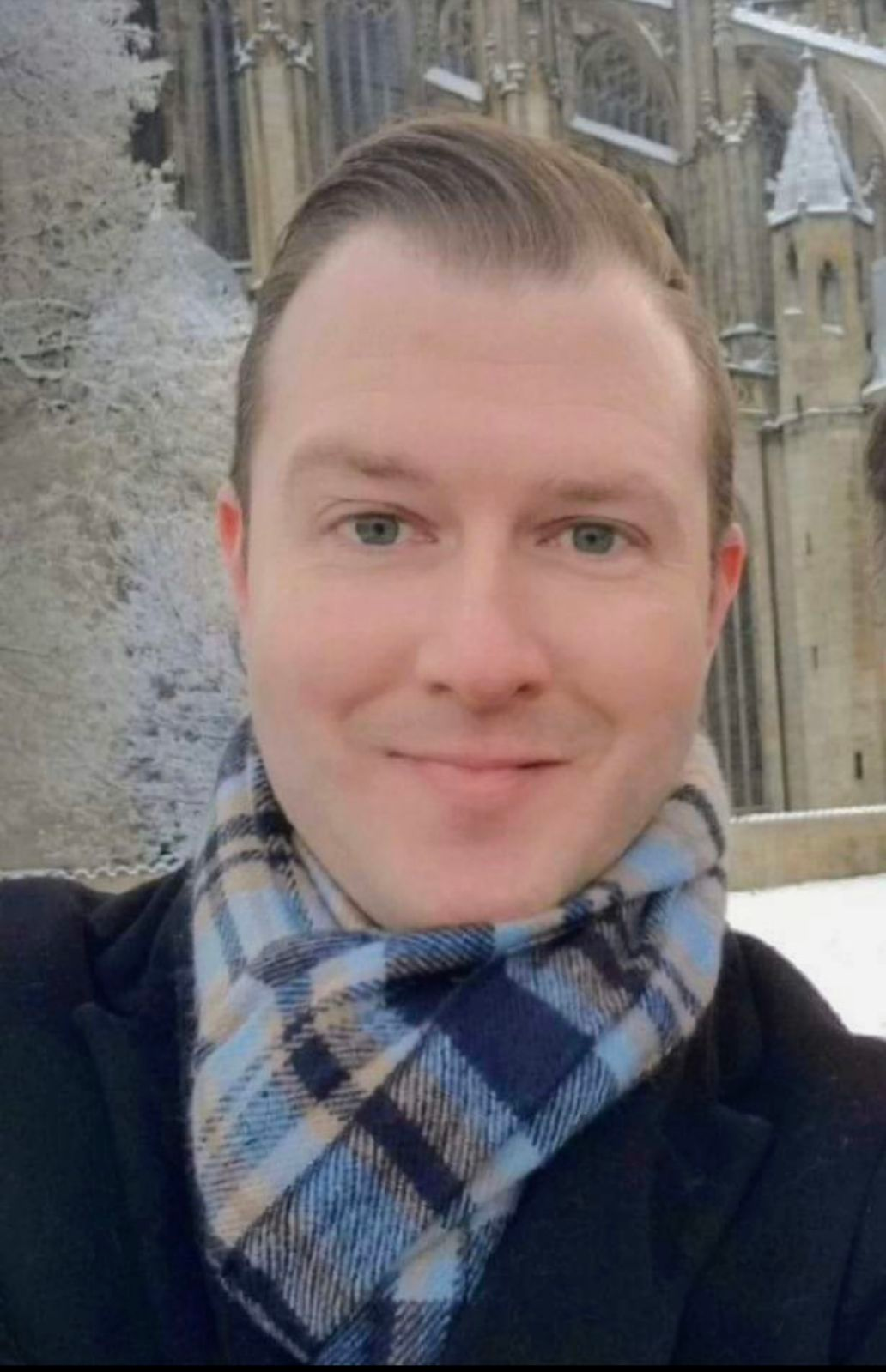 A man wearing a plaid scarf is smiling in front of a building.