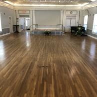 Empty ballroom with polished wooden floor, white walls, and a raised stage at the far end.