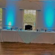 Event table with white cloth, blue uplighting, and a window-centered backdrop in a room with wood flooring