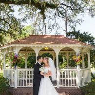 Bride and groom kissing in a white gazebo with a red roof, surrounded by trees and flowers.