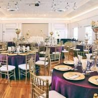 Elegant banquet hall set with round tables, gold chairs, and purple tablecloths under bright chandeliers
