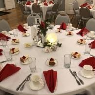 Round banquet table set with red napkins, cups, dessert plates, and a glowing centerpiece in a dining hall