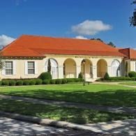 Single-story cream building with red tile roof and arched walkway, beside a green lawn under a blue sky
