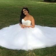 Woman in a white strapless ball gown sitting on grass outdoors, smiling at the camera.