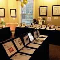 Framed artwork and documents displayed on black-draped tables in a gallery room with yellow walls and windows.