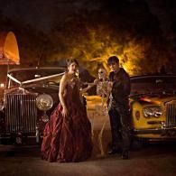 Two people in formal attire pose between classic cars at night, lit by warm orange lights.