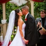 Bride and groom kiss at an outdoor wedding ceremony while an officiant holds a microphone nearby