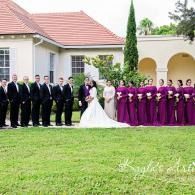 Wedding party posing on a lawn, bride in white with attendants in purple and groomsmen in black suits.