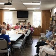People seated around a long table in a bright meeting room with bookshelves and a wall-mounted TV.