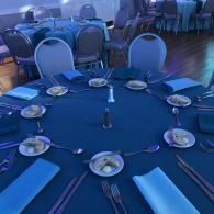 Blue-clothed round banquet table set with plates, glasses, napkins, and silverware in a dim event hall