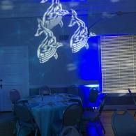 Blue-lit event space with tables and chairs, featuring a projected abstract white figure on the wall.