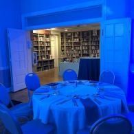 Blue-lit banquet room with round tables and chairs, open doorway to a library in the background