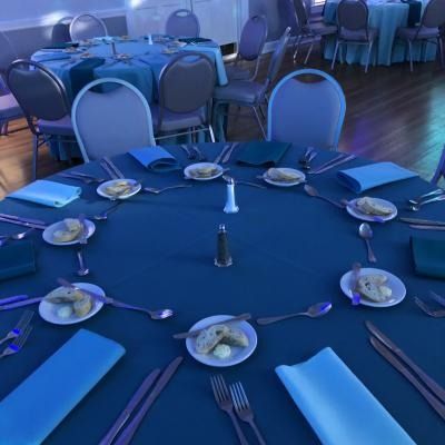 Blue-lit banquet table set with plates, forks, spoons, and folded napkins in a dining hall