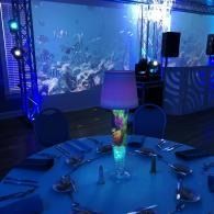 Blue-lit banquet table with a glowing lamp, set before a large aquarium in a dim room.