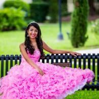 Woman in a bright pink ruffled gown sitting on a park bench, smiling in a green garden setting