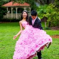 Couple in formal wear smiling in a grassy garden, the young woman being picked up in her pink ruffled dress.