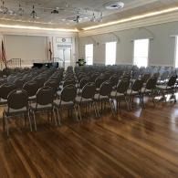 Rows of black chairs in a bright banquet hall with wood flooring and a stage at the front