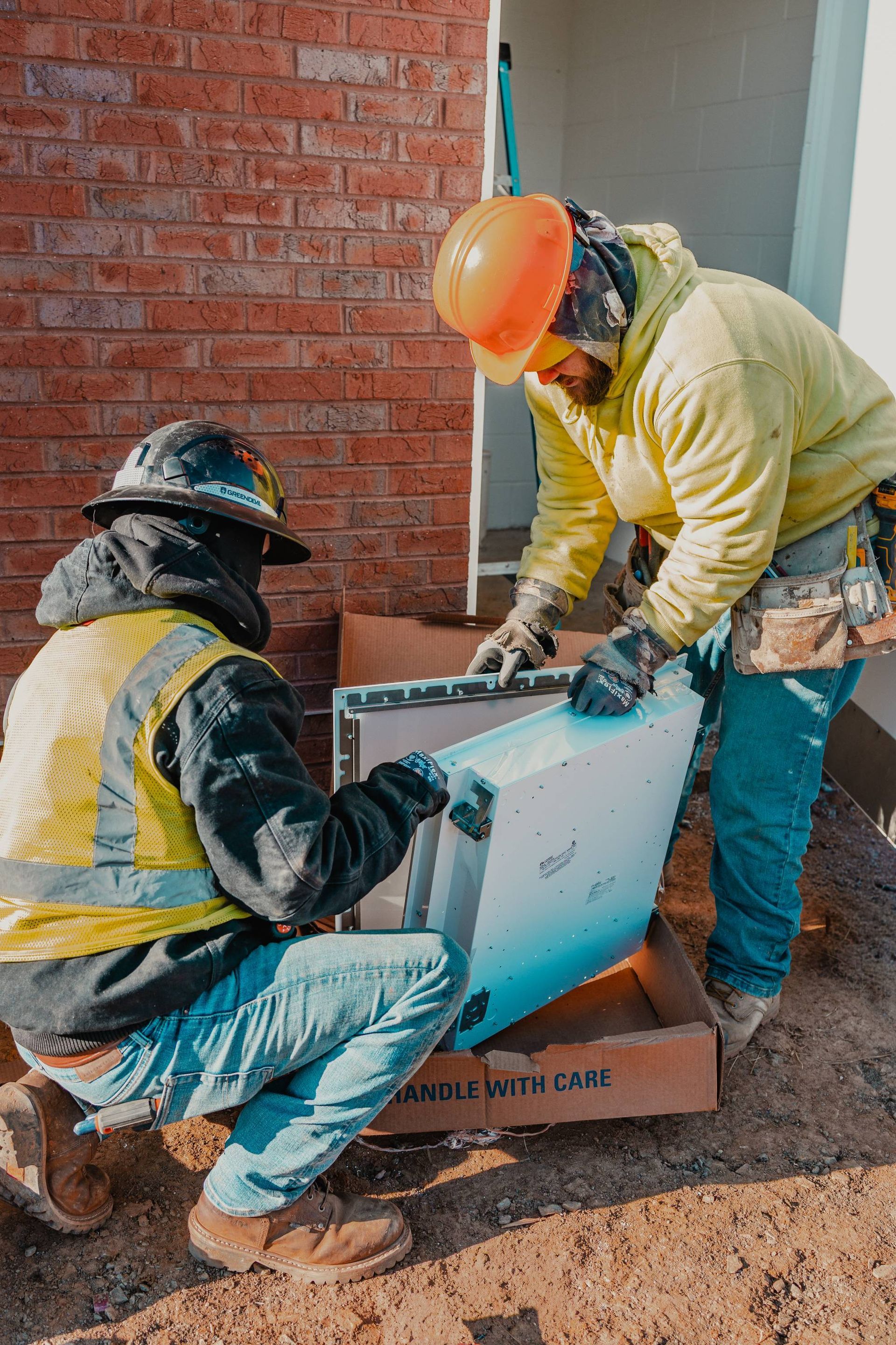 Two construction workers installing a white box. One kneels, the other stands. Brick wall in background.