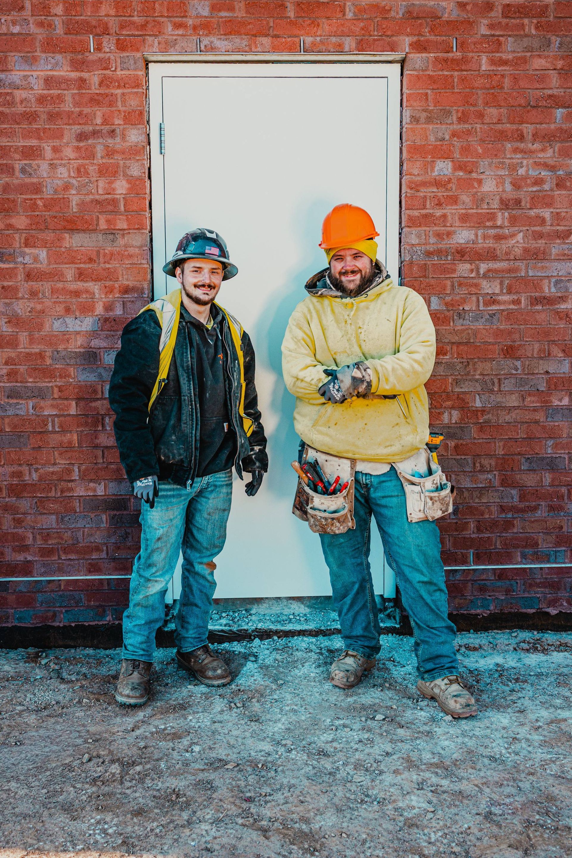 Two construction workers in front of a white door on a brick building. One in yellow, one in black, both wearing safety gear.