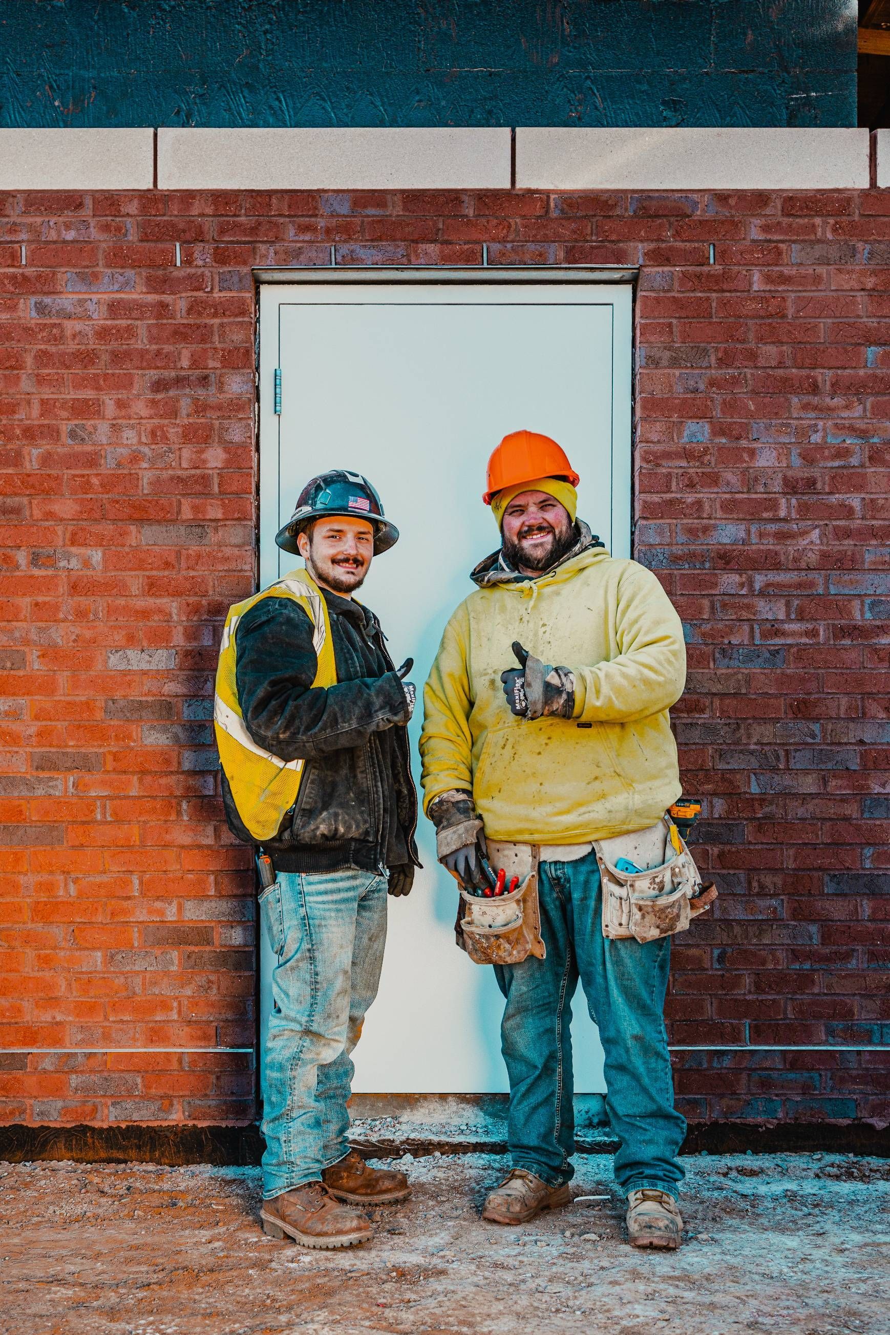 Two construction workers stand in front of a white door on a brick building, giving thumbs up.