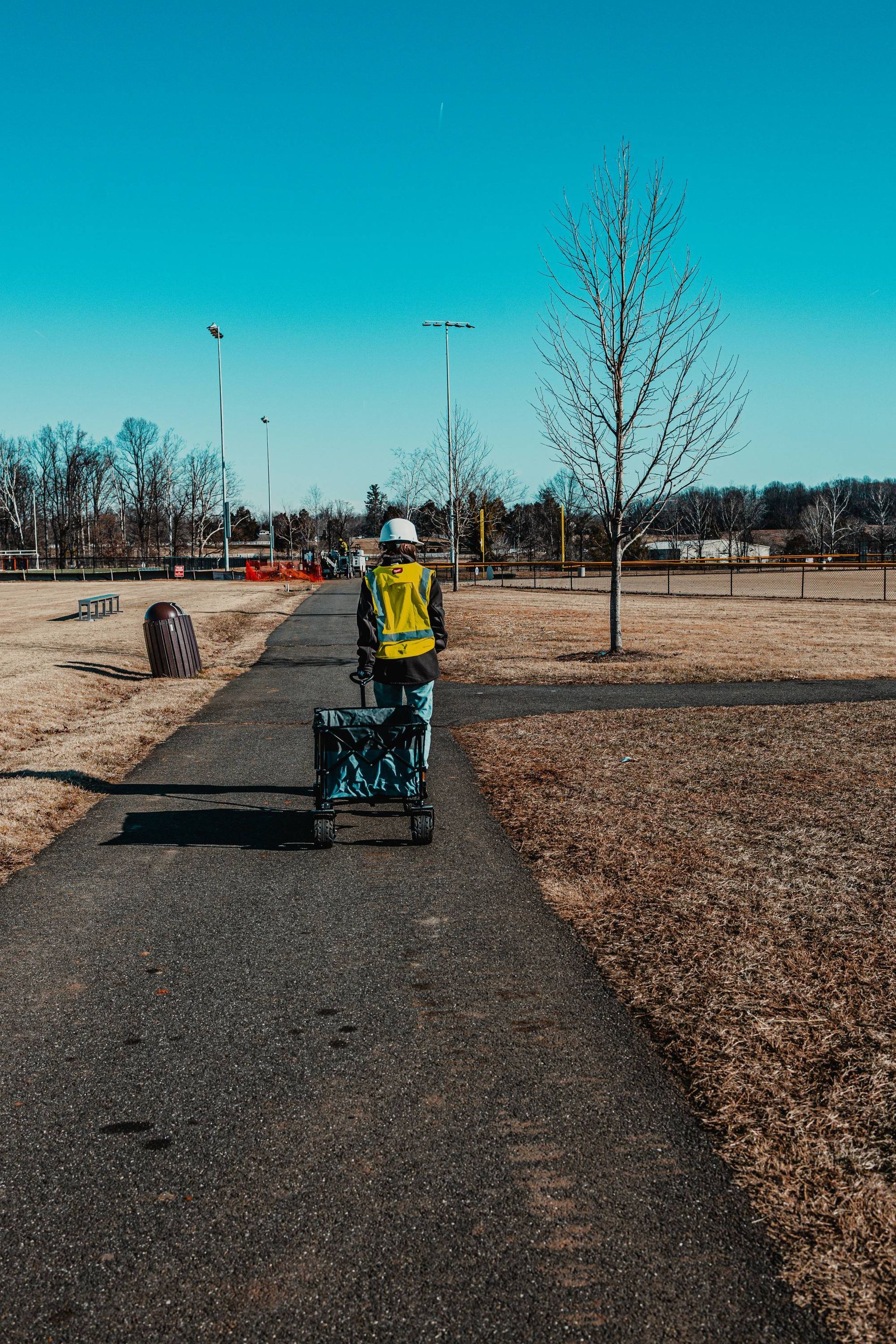 Person wearing a safety vest pulls a cart along a paved path in a park on a sunny day.