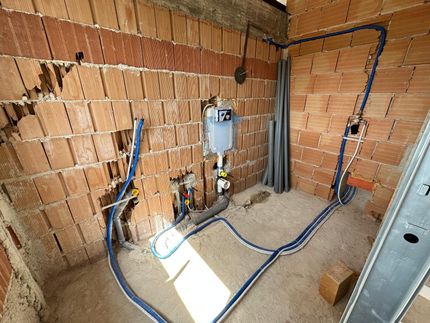 Locale tecnico non finito con pareti di mattoni a vista, tubazioni blu, uno scaldabagno e quadri elettrici.