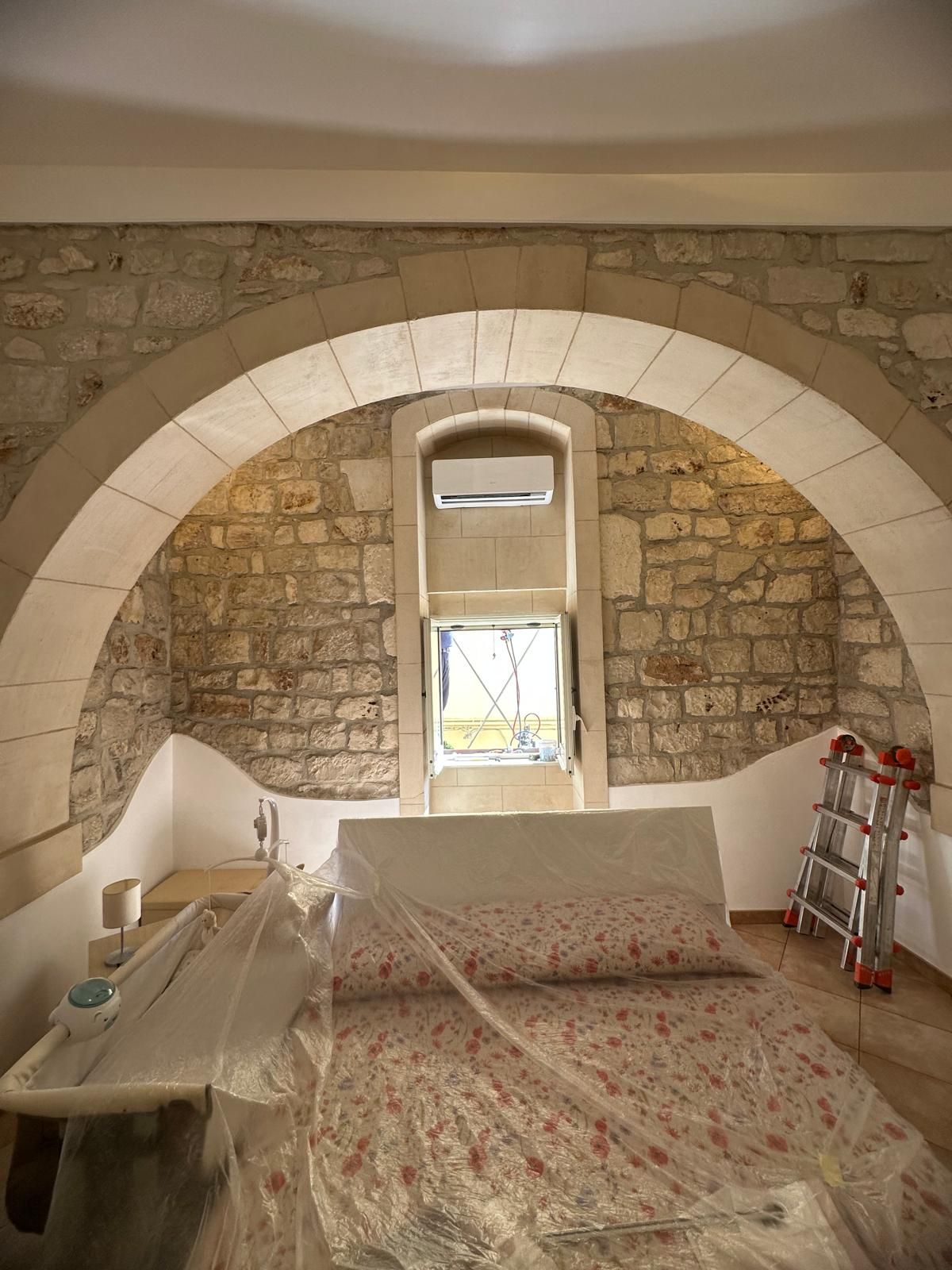 Camera da letto con parete in pietra a vista e arco bianco, letto al centro, finestrella e scala a destra