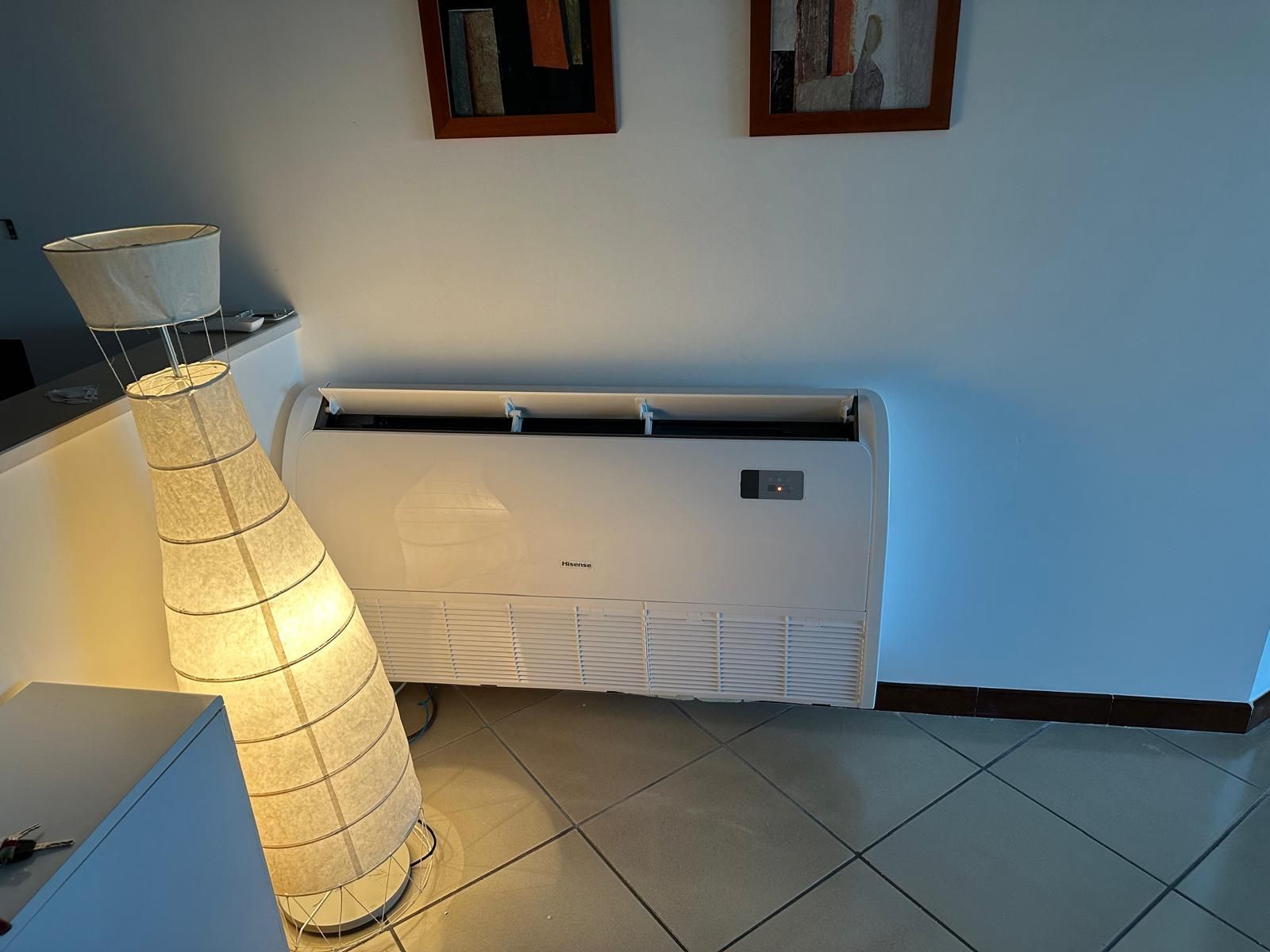 Lampada da terra accanto a un termosifone bianco a parete, in una stanza piastrellata con quadri incorniciati appesi sopra.
