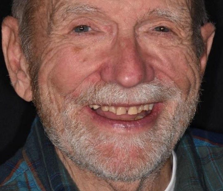 Older man with a gray beard smiling, showing teeth, wearing a plaid shirt.