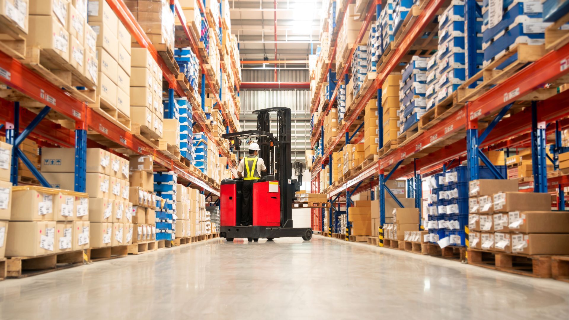 A warehouse worker operates a red forklift in a narrow aisle between tall industrial shelving stacked with boxes.
