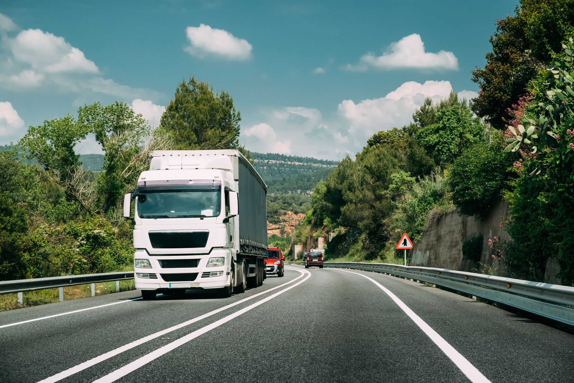 A white semi-truck driving on a paved highway surrounded by lush green trees under a bright blue sky.