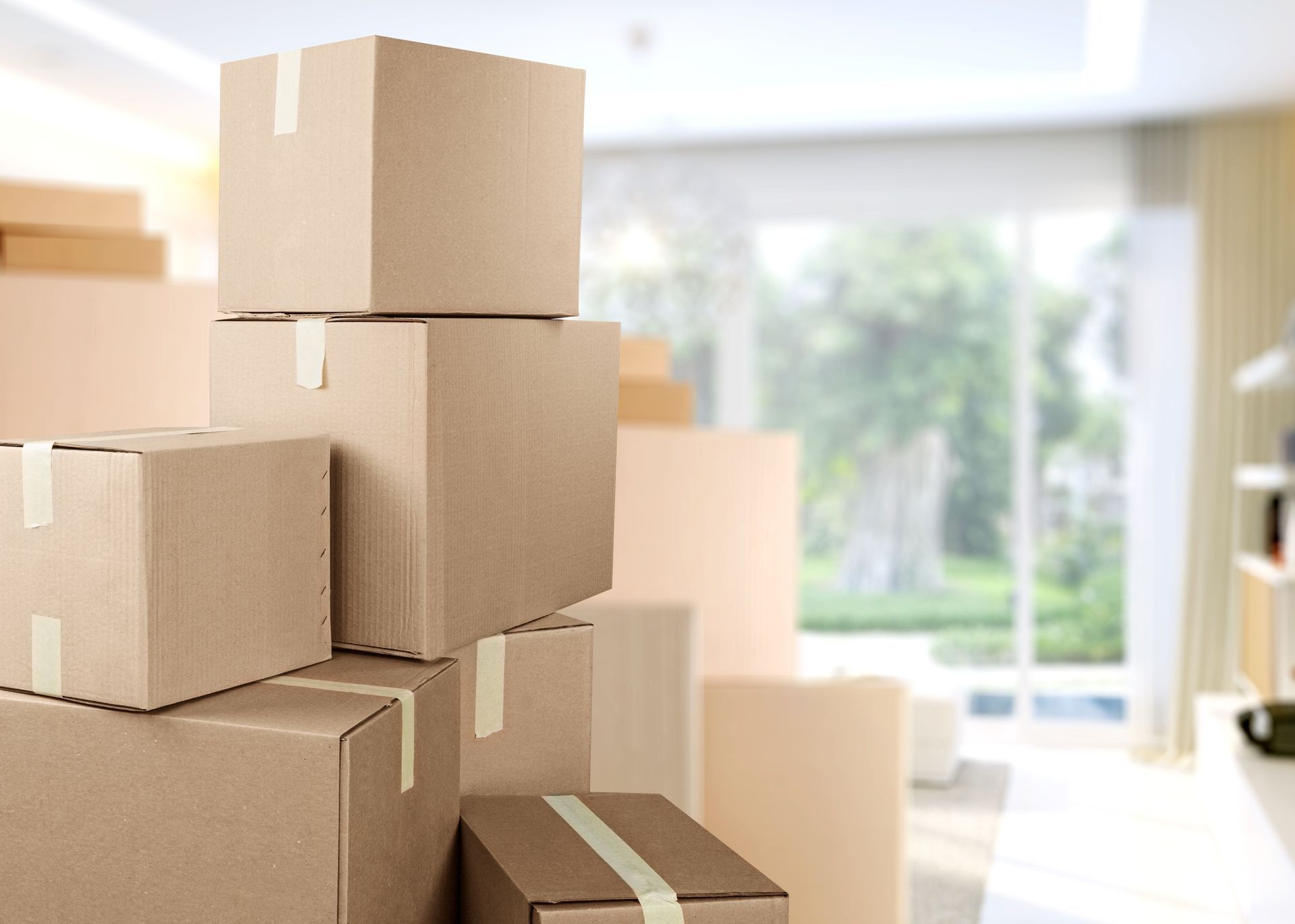 A stack of cardboard moving boxes in a blurred, sunlit home interior.