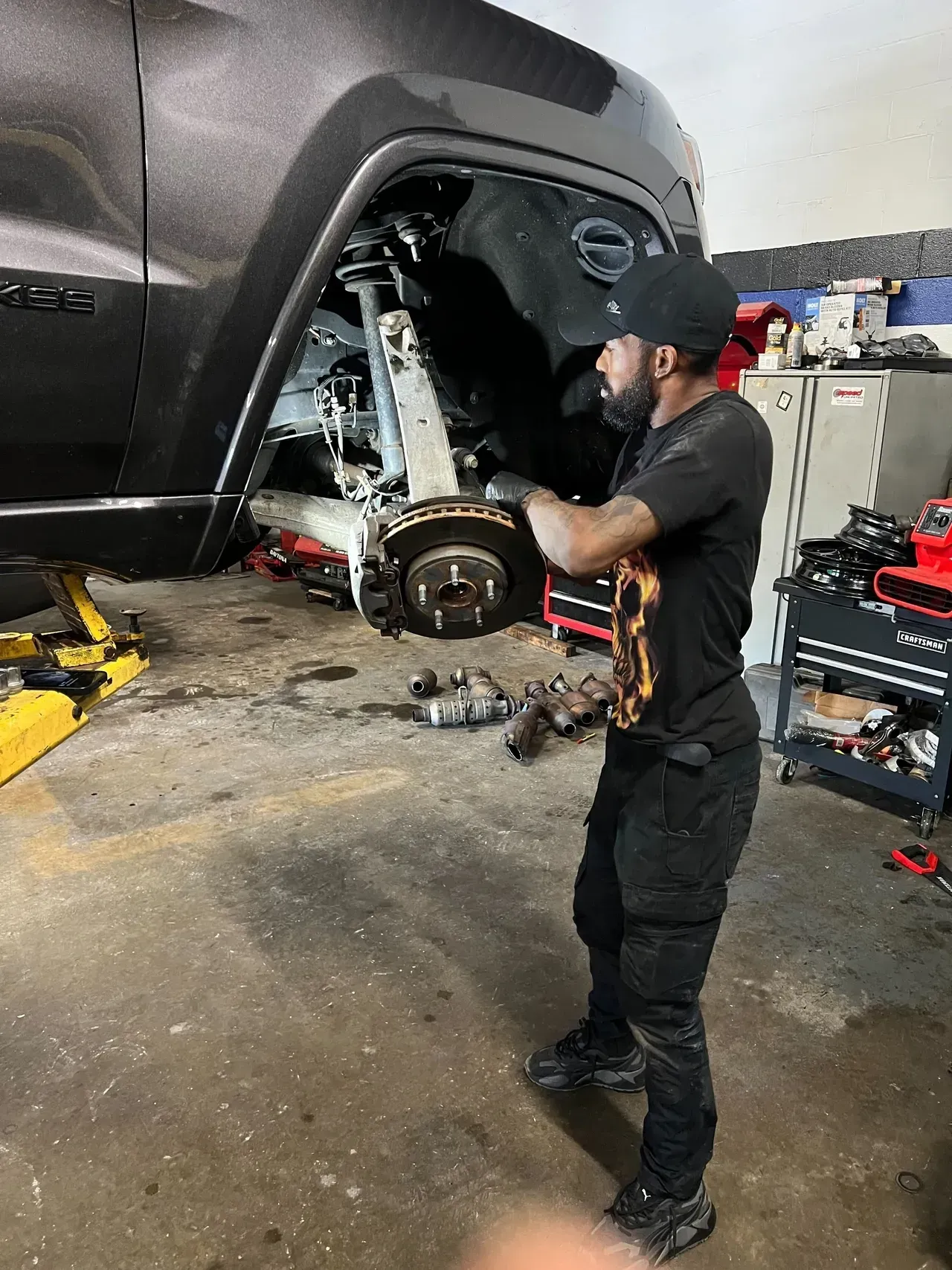 Mechanic working on the front wheel of a car in a garage. Wearing a black cap and gloves. | Ashten's Total Auto Care