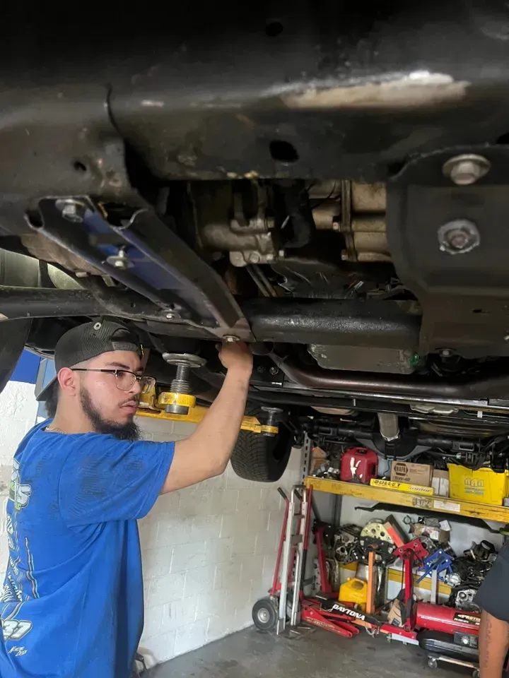 Mechanic working under a vehicle in a garage. Wearing blue shirt and hat, reaching up. | Ashten's Total Auto Care