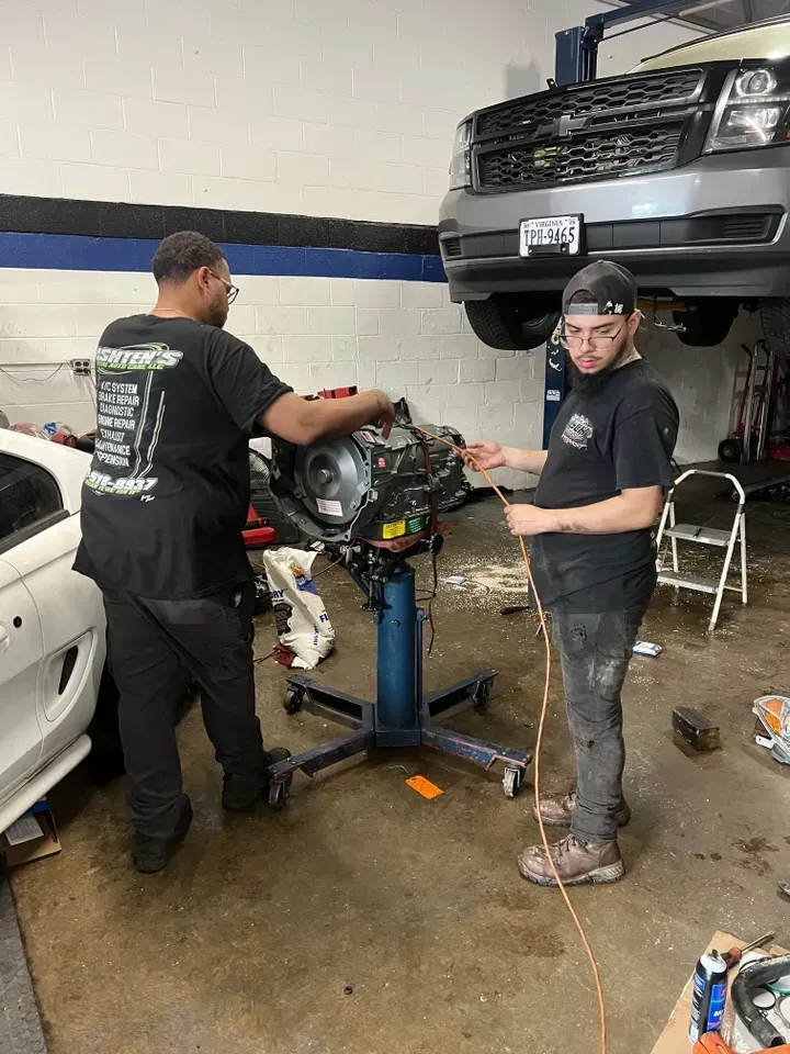 Two mechanics working on a car engine in a garage. One holds a wire, the other works on the engine. A car is lifted in the background. | Ashten's Total Auto Care