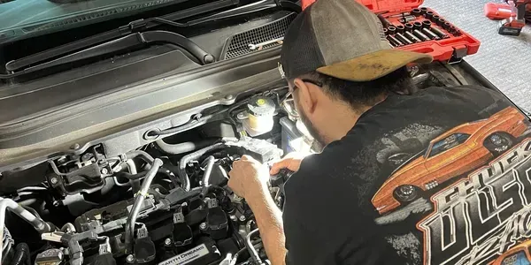 Person working on a car engine. Wearing a hat and a shirt with a car design. Tools are visible. | Ashten's Total Auto Care