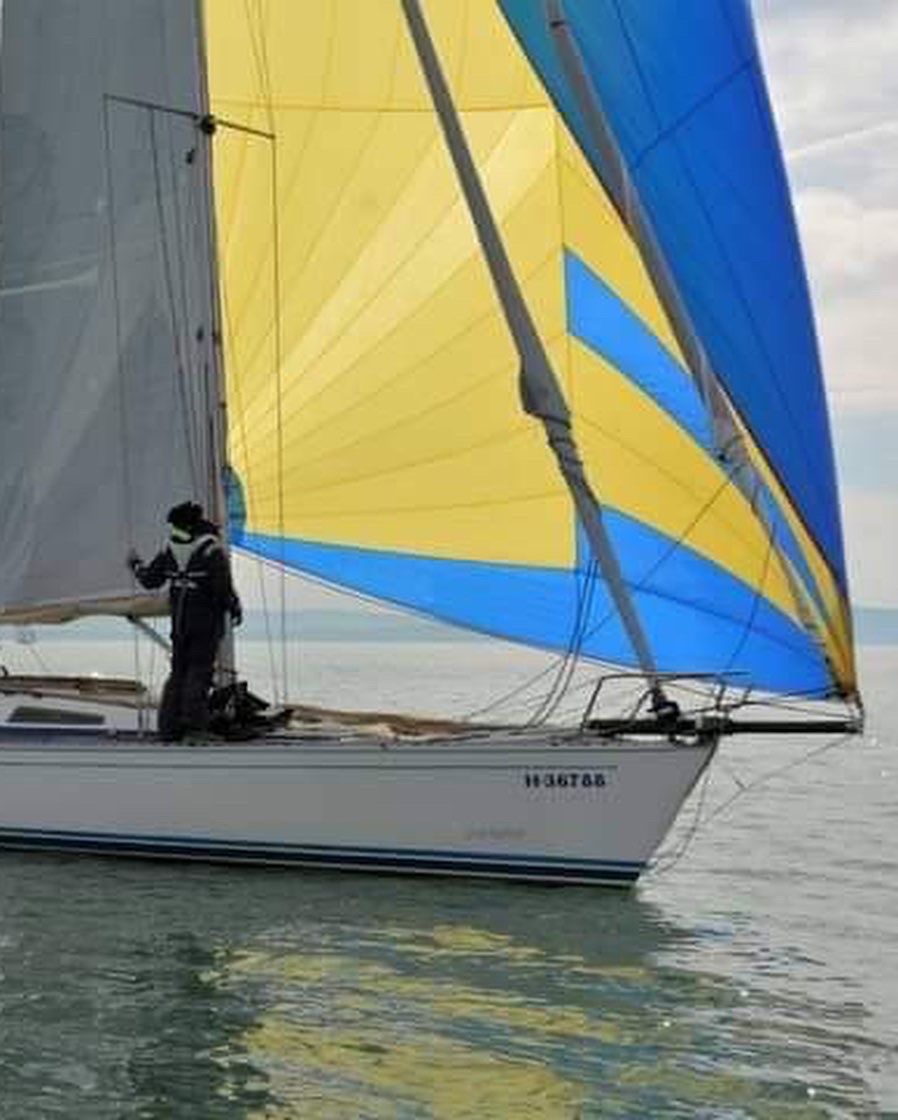 Sailboat with a yellow and blue sail. A person on deck adjusts something. Water.