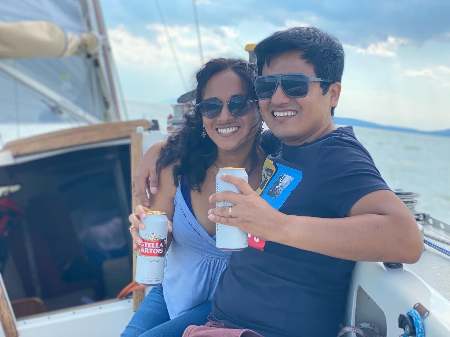 A couple on a sailboat, smiling and holding beers. Blue sky and water in the background.