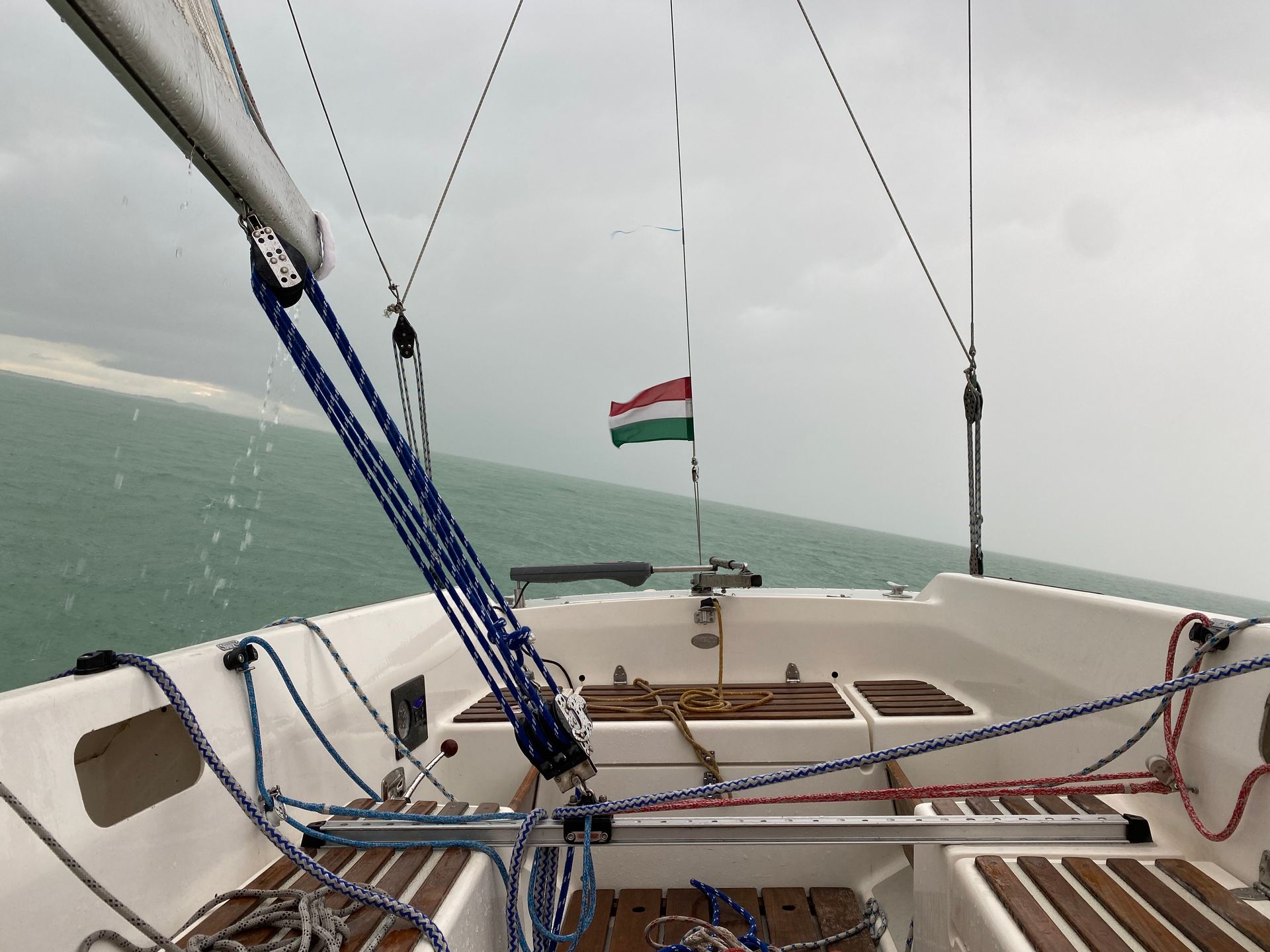 Sailboat deck with Hungarian flag flying, looking towards hazy horizon over green water.