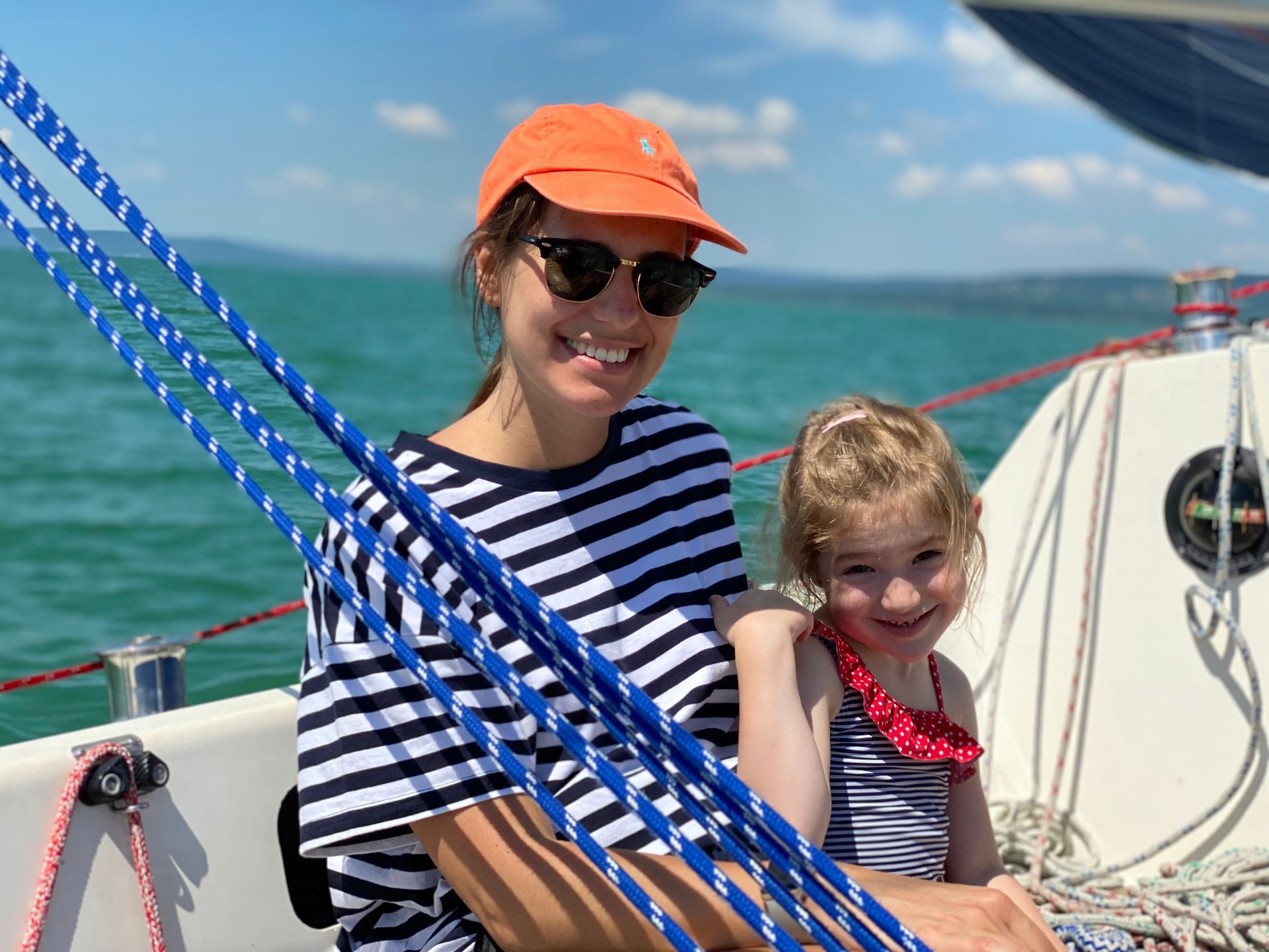 Woman and child smiling on a sailboat, wearing sunglasses and striped shirts, blue ropes, sunny day.