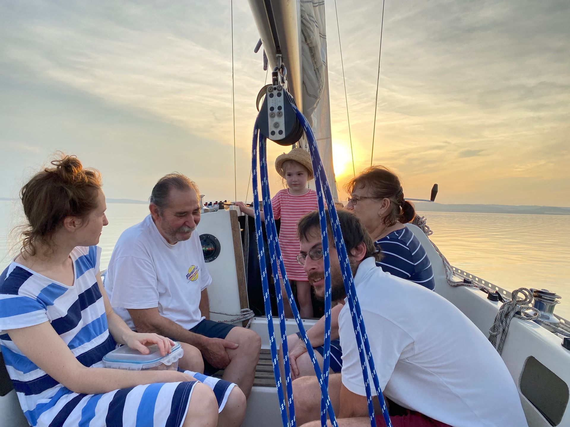 Family on a sailboat at sunset. People smiling. Sky is orange and cloudy.