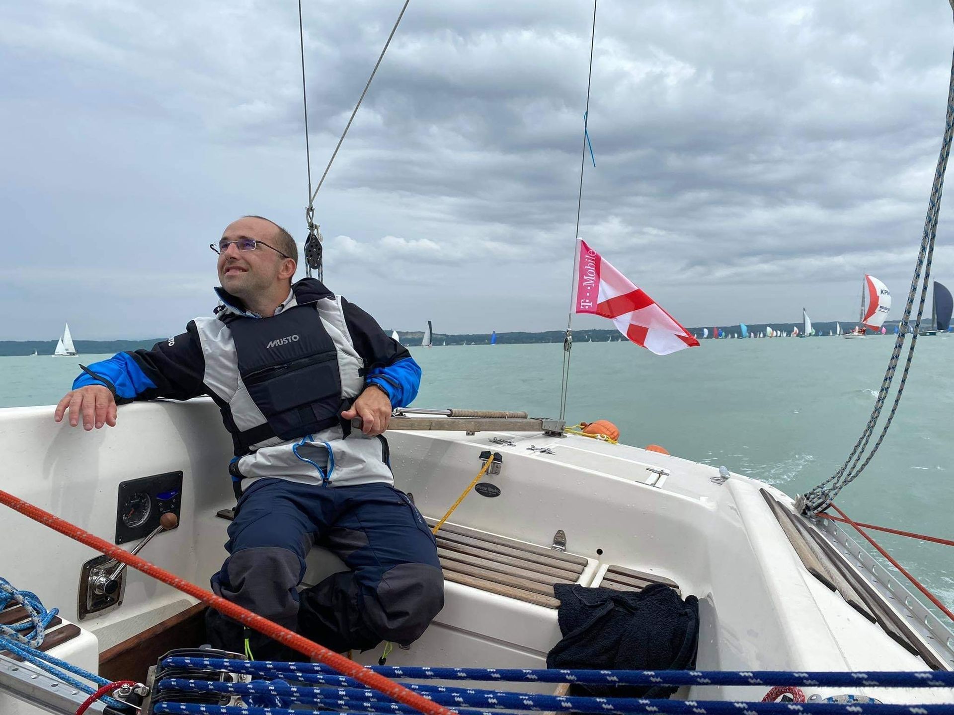 Man on a sailboat, looking up with a smile, on the water with other sailboats. Cloudy sky.